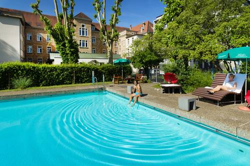 Quelle: Erzbischöfliches Seelsorgeamt Freiburg - Bildungshäuser und Familienferienstätten Mann sitzt am Beckenrand des Außenschwimmbades und Frau liegt auf einer Liege im Hintergrund das Bildungshaus St. Bernhard