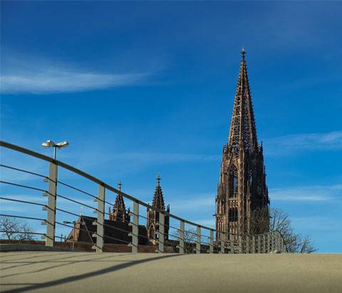 Quelle: Pixabay Im Vordergrund ist eine Brücke mit Geländer und im Hintergrund der Turm des Freiburger Münsters vor blauem Himmel und einzelnen Wolken.